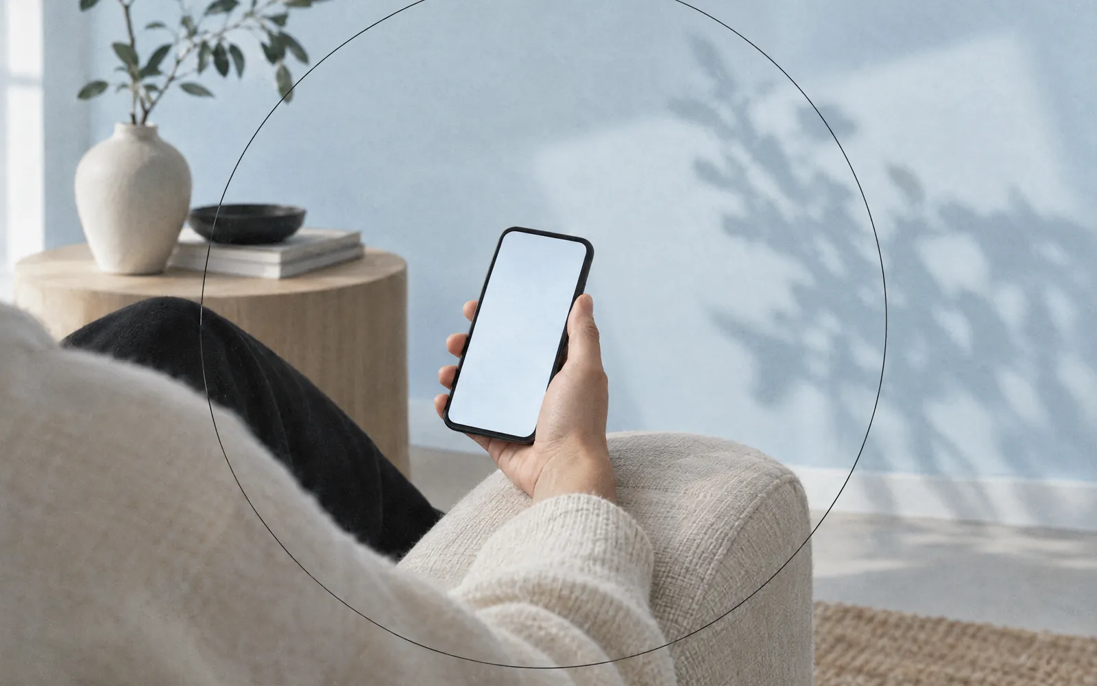 Eine Hand hält ein Smartphone mit leerem Bildschirm auf der Sofalehne, hellblau getönter Wohnraum.